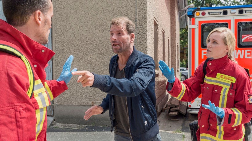 Das Bild zeigt eine angespannte Situation in einem Rettungswagen. Zwei Rettungskräfte in roten Uniformen mit reflektierenden Streifen stehen einem Mann gegenüber, der scheinbar aggressiv oder unkooperativ wirkt. Der Mann wird von einem der Rettungskräfte an den Schultern gehalten, vermutlich um ihn zu beruhigen oder zurückzuhalten. Die andere Rettungskraft, eine Frau, schaut aufmerksam und besorgt auf die Situation.