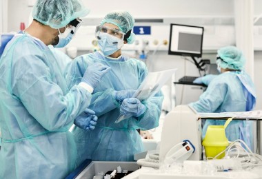 Foto von einer Ärztin und einem Arzt, die in Schutzkleidung auf einer Intensivstation stehen und sich beratschlagen.