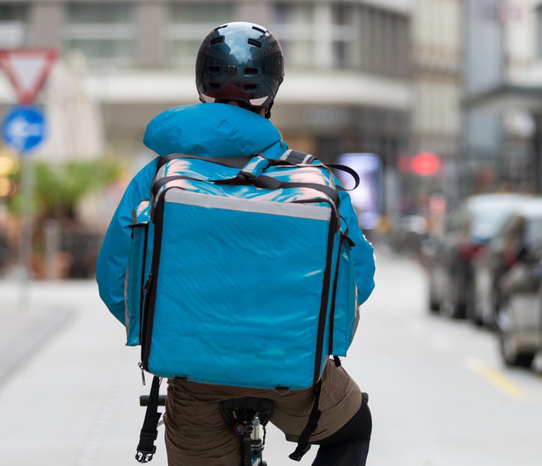 Die Person trägt einen dunklen Helm, sowie eine Jacke und einen Lieferdienst-Rucksack im gleichen hellblauen Farbton. 