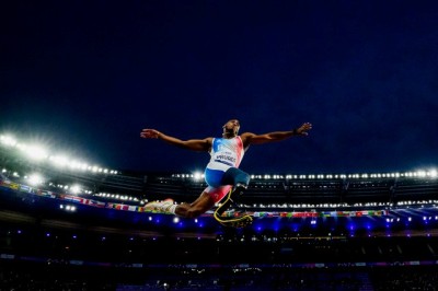 Nächtliche Szene: Ein Weitspringer mit Beinprothese springt vor und über dem Betrachter vorbei. Die Bewegungen sind eingefroren, die Arme weit auseinander, das Stadion im Hintergrund, Flutlichtern tauchen alles in eine fast surreale Athmosphäre.